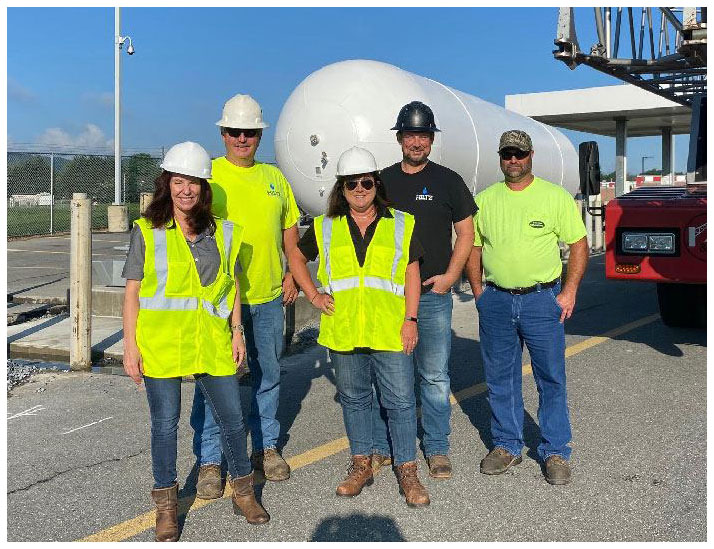 Five propane workers in reflective safety vests and hardhats smile in front of a propane transport tank