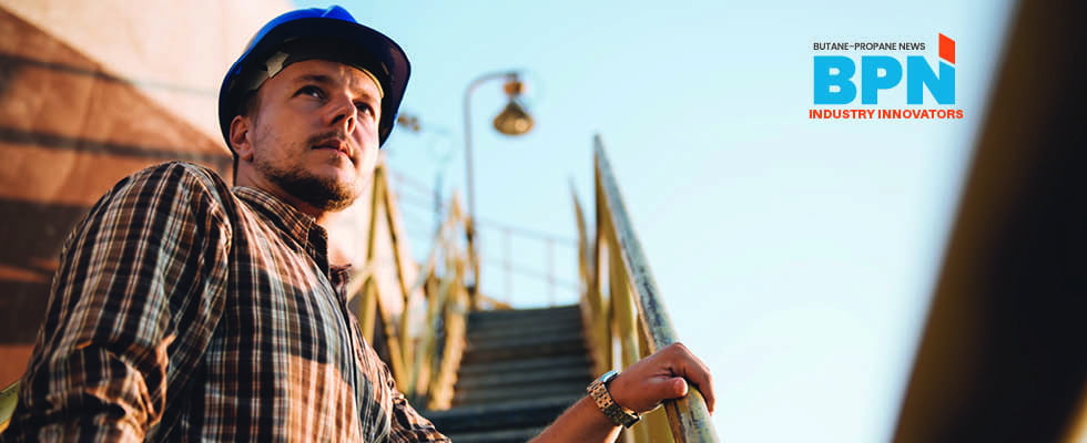 A male worker in a plaid shirt and blue hard hat stands at the bottom of yellow stairs at an industrial site