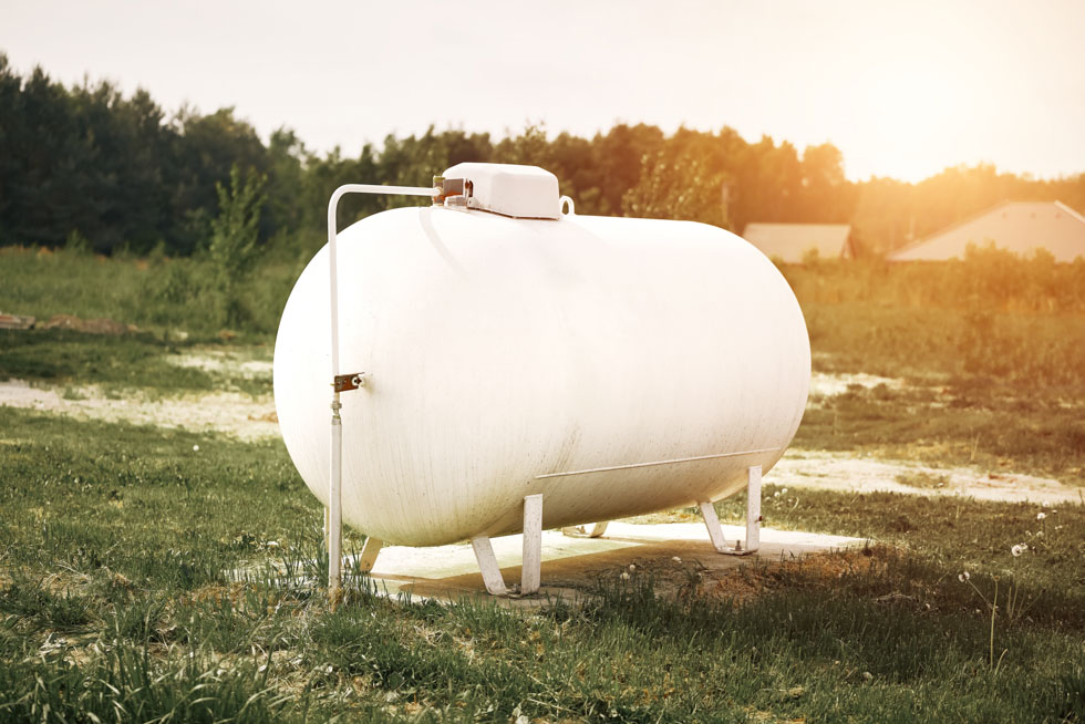 A white propane tank sits in a green field with a house and trees in the distance, representing the 2026 state of the propane industry
