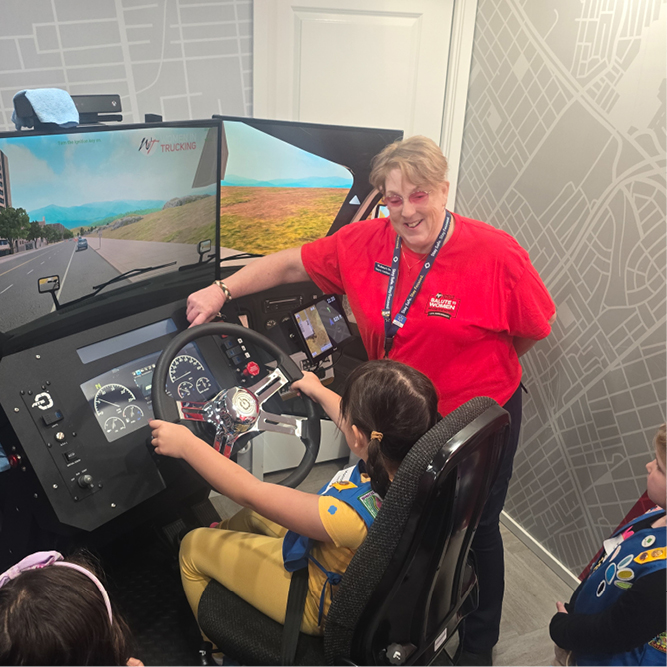 A Girl Scout sits behind the wheel of a driving simulator while a female truck driver stands beside her at the Trucks Are for Girls event