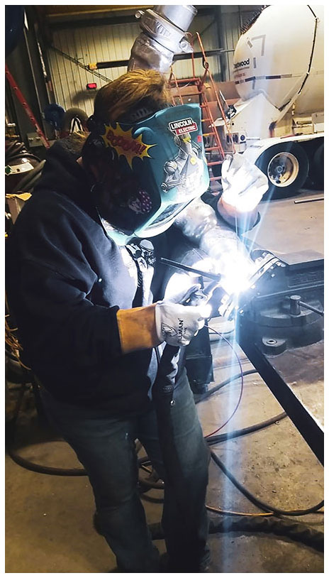 Cathy Maloy works on a welding project while wearing gloves and heavy-duty protective face gear covered in personalized stickers.