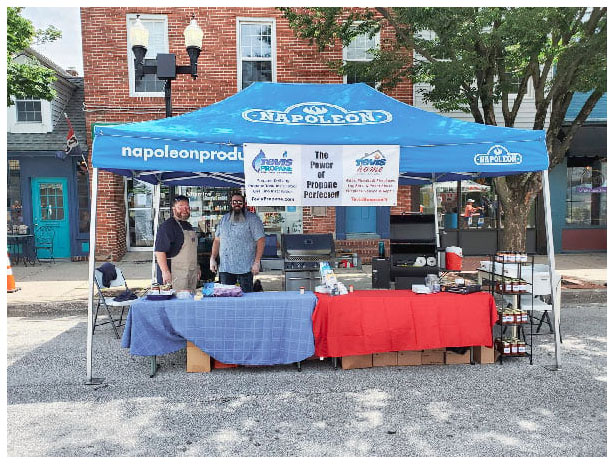 Two men run a Tevis Propane street fair booth offering food cooked on a propane-powered grill.