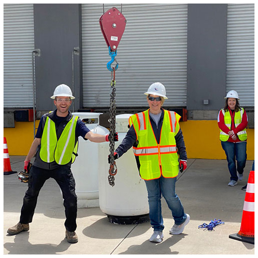 Two employees in personal protective equipment vests, hard hats and gloves pose with the hook of a crane