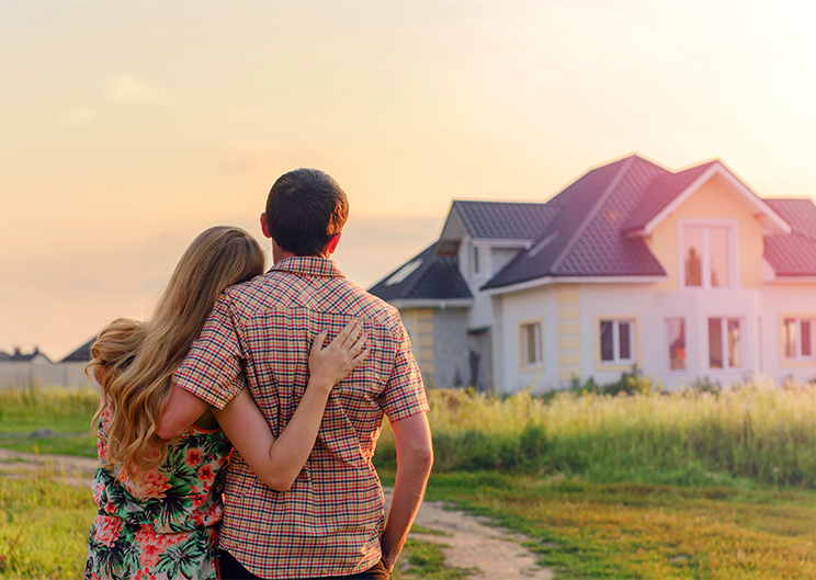 A couple stands with their back turned to the camera, gazing into the distance at a house lit by the setting sun.