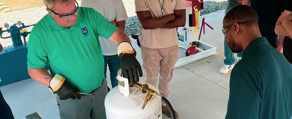 An educator in a green shirt and safety glasses and gloves shows students how to work with a propane tank.