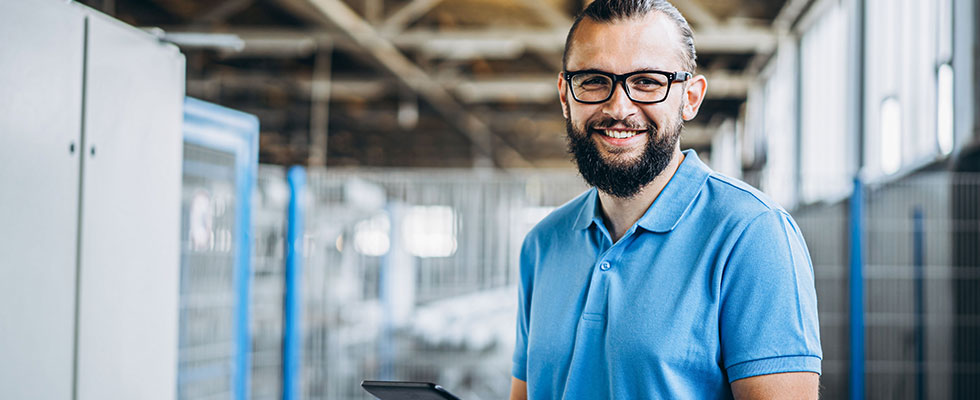 A younger man with a beard, glasses and blue collard shirt works from an iPad at work