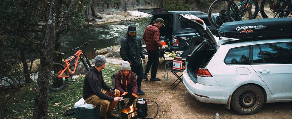 A woman and three men in winter camping gear gather around a campfire fueled by a Gas Growler propane cylinder