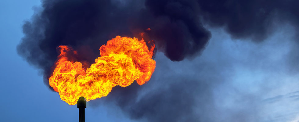 Fire burns and smoke billows out of a factory smokestack against a blue sky