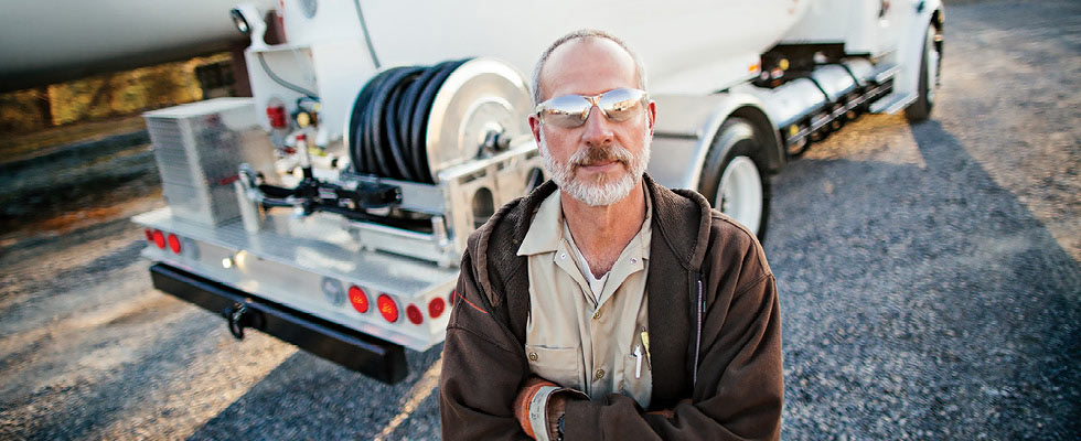 A bobtail driver in protective eyewear and gloves stands in front of a bobtail truck with his arms crossed