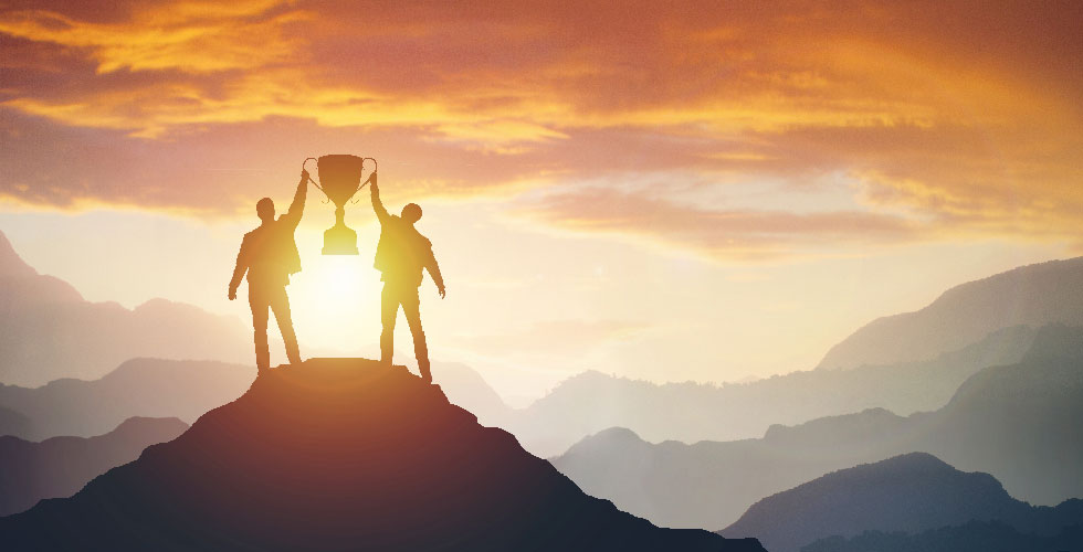 Two men stand on a mountain peak in front of a sunset, holding up a large trophy between them.
