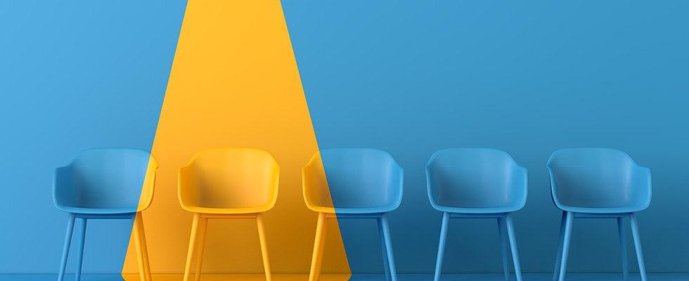 A row of five chairs in front of a blue background. Four chairs are blue — one chair is yellow with a yellow spotlight shining down on it