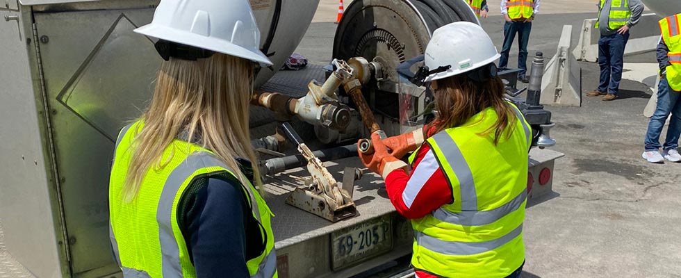 Two women in personal protective equipment vests, hard hats and gloves train on the equipment on the back of a bobtail truck