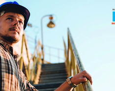 A male worker in a plaid shirt and blue hard hat stands at the bottom of yellow stairs at an industrial site