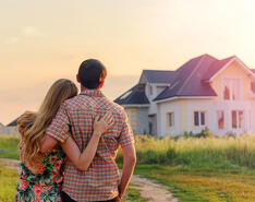 A couple stands with their back turned to the camera, gazing into the distance at a house lit by the setting sun.