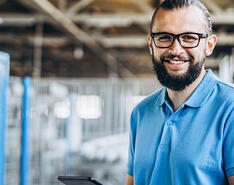 A younger man with a beard, glasses and blue collard shirt works from an iPad at work