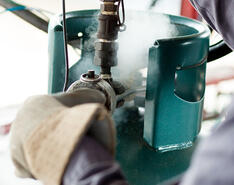 A person wearing protective gloves works with the valve of a teal propane tank while gas releases around the valve