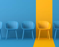 Five chairs in a row (three blue, one yellow, then one blue) on a blue background with a yellow spotlight on the yellow chair