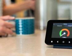 A smart gas meter sits on the counter with a hand holding a coffee cup out of focus in the background