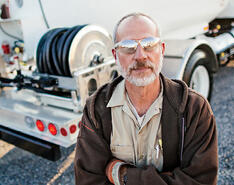 A bobtail driver in protective eyewear and gloves stands in front of a bobtail truck with his arms crossed