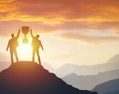 Two men stand on a mountain peak in front of a sunset, holding up a large trophy between them.