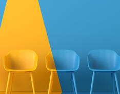 A row of five chairs in front of a blue background. Four chairs are blue — one chair is yellow with a yellow spotlight shining down on it