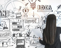 A woman stands drawing on a whiteboard various diagrams and charts representing marketing planning.