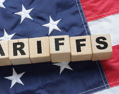 Block lettering spelling out tariffs in front of the U.S. flag