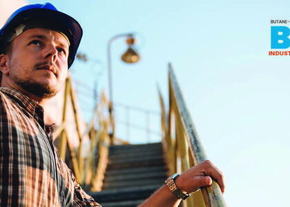 A male worker in a plaid shirt and blue hard hat stands at the bottom of yellow stairs at an industrial site