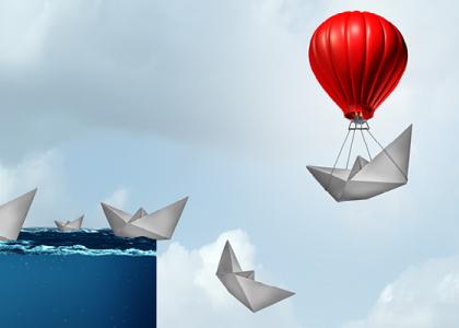 Paper boats fall over the edge of a cliff of water while a red hot air balloon carries one away to safety