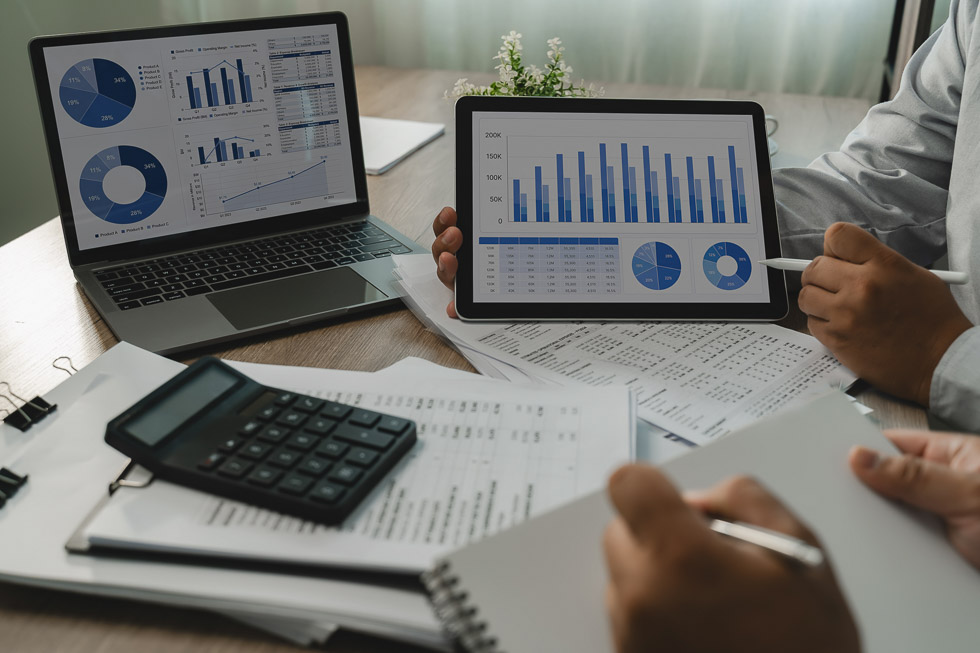 Business team reviews charts on a laptop and tablet beside printed reports and calculator, comparing figures and dashboard data for business analysis, reporting and managing financial risk .